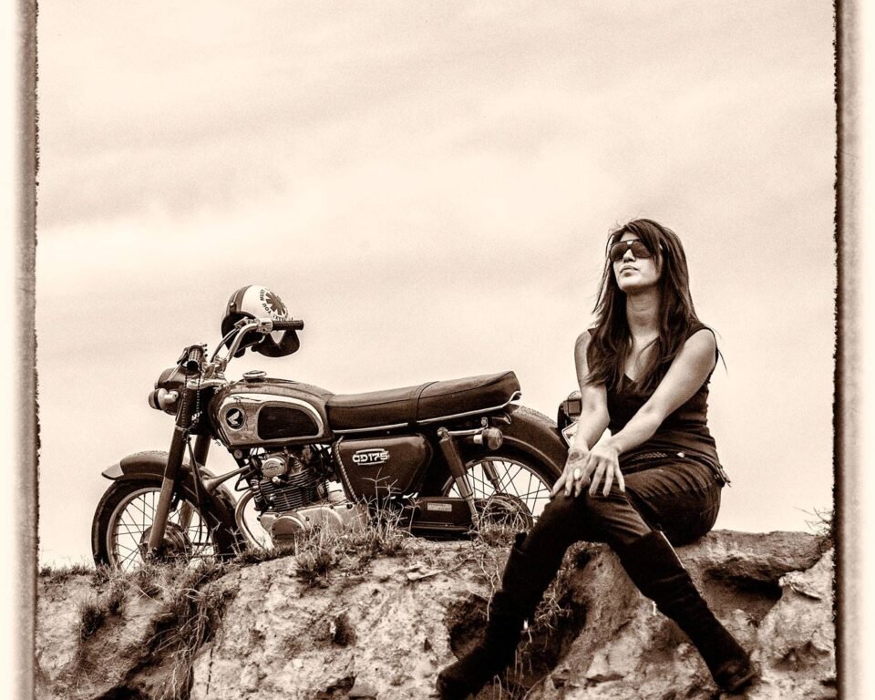 Wide shot, sepia-toned editorial image of a woman in sunglasses and dark clothing sitting on a rocky outcrop next to a vintage motorcycle under a wide sky.