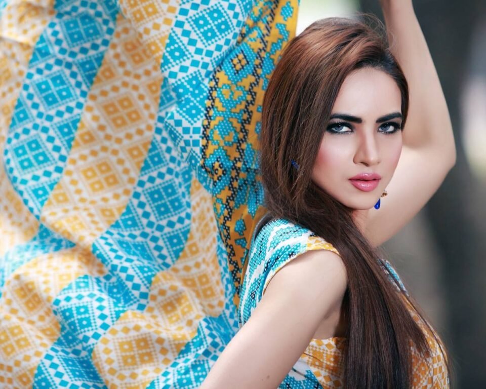 Close-up editorial portrait of a woman with long brown hair, bright makeup, posing with a large swath of flowing fabric in a blue and yellow geometric print.