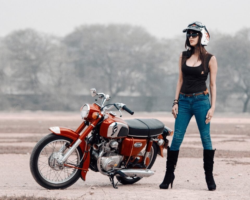 Full-length editorial photograph of model Sania Baig Mirza standing next to a vintage red and chrome motorcycle, wearing a black tank top, jeans, black boots, and a helmet pushed up over sunglasses. Editorial fashion photography by AHSANZ.