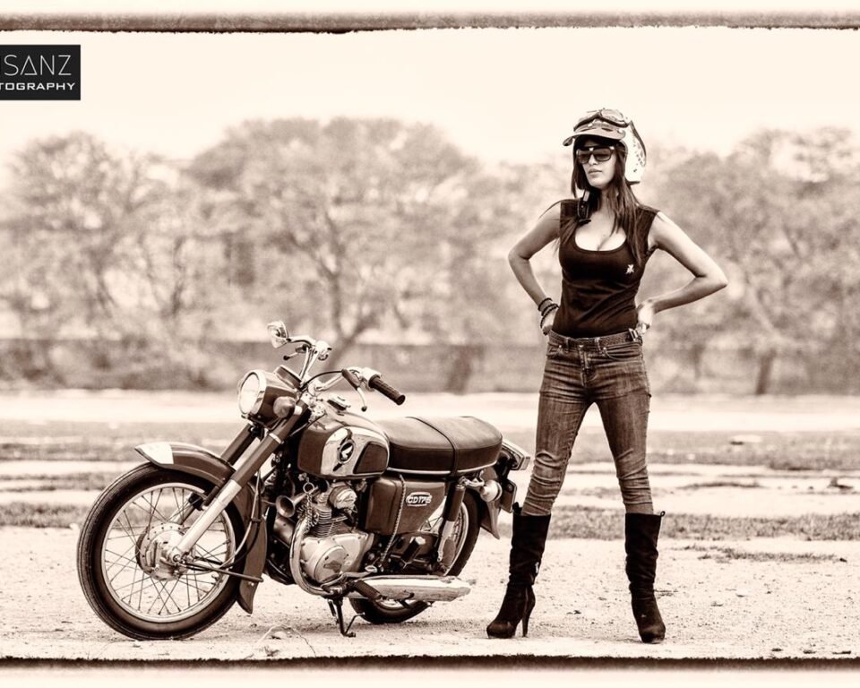 Sepia-toned (brown and white) full-length editorial photograph of a model (Sania Baig Mirza) wearing a helmet, sunglasses, black top, and jeans, standing next to a vintage motorcycle. Artistic sepia editorial fashion photography by AHSANZ.