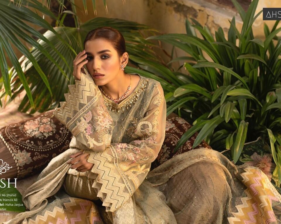 Editorial fashion photograph for the 'LUSH' campaign featuring model Nafisa Janjua seated among lush greenery and patterned pillows, wearing a gold and beige embroidered traditional Pakistani outfit (wardrobe by LUSH). Fashion and jewelry photography by AHSANZ.