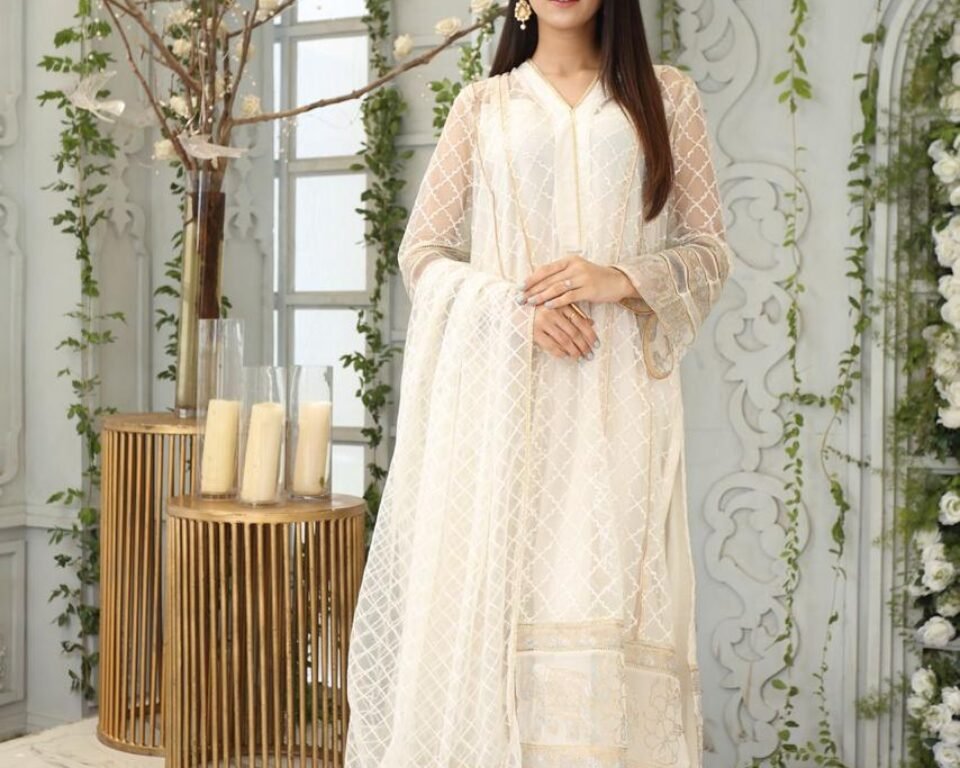 Full-length editorial fashion photograph of a model standing in a bright, white, ornate room decorated with flowers and greenery, wearing an elegant white and gold embroidered traditional Pakistani suit with a sheer dupatta. Fashion photography by AHSANZ.