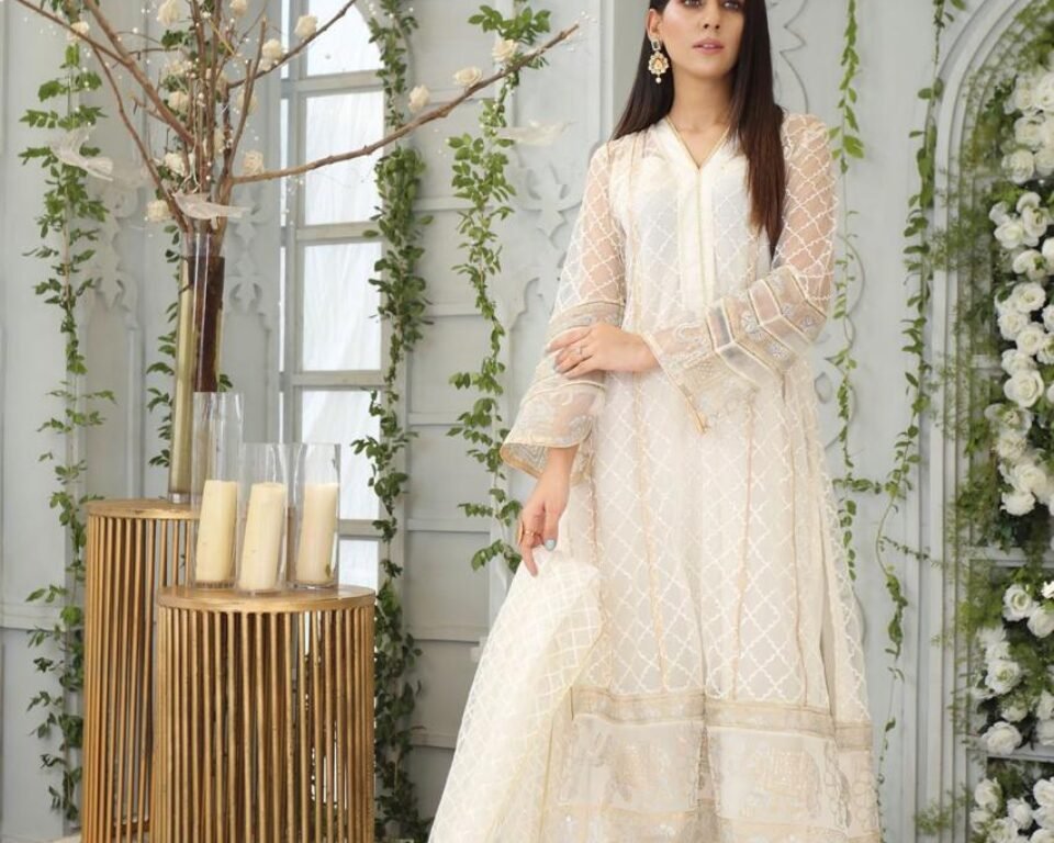 Full-length editorial fashion photograph of a model in a bright white room with vine decoration, wearing an elegant white sheer kurta and trousers with gold trimming, and crystal-embellished heels. Fashion photography by AHSANZ.