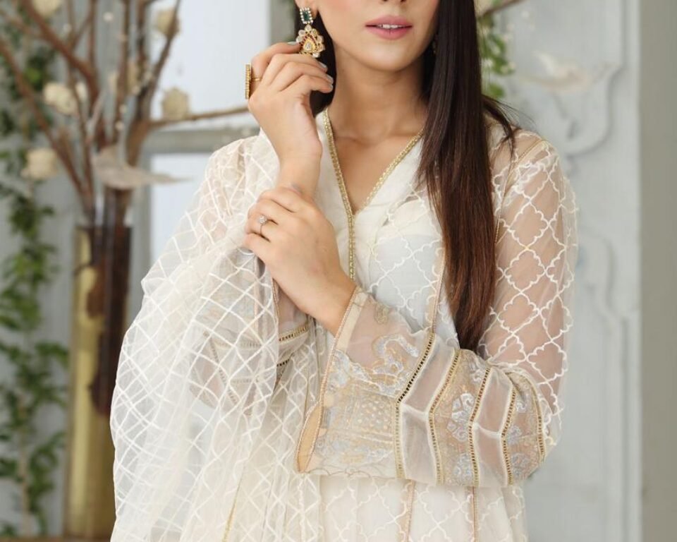 Close-up editorial fashion portrait of a model in a bright white room, wearing an elegant white sheer kurta with gold embroidery on the sleeves, holding her hand near her face to show a ring and traditional jhumka earrings. Fashion and jewelry photography by AHSANZ.