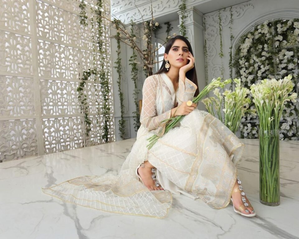 Editorial fashion portrait of a model sitting on a white marble floor, holding a bouquet of white flowers, wearing an elegant white and gold embroidered traditional Pakistani suit with a sheer dupatta. Fashion photography by AHSANZ.