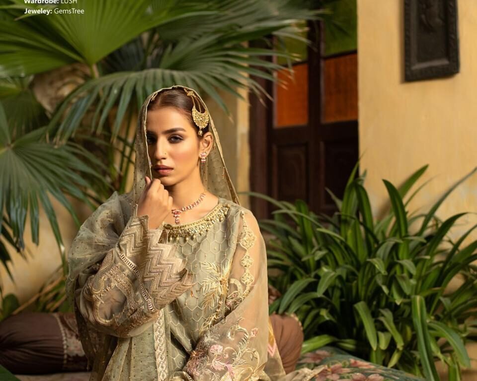 Vertical editorial fashion portrait for the 'LUSH' campaign featuring model Nafisa Janjua seated with a dupatta over her head, wearing a gold and pale green heavily embroidered traditional Pakistani outfit (Wardrobe: LUSH, Jewelry: Gemstree). Fashion and jewelry photography by AHSANZ.