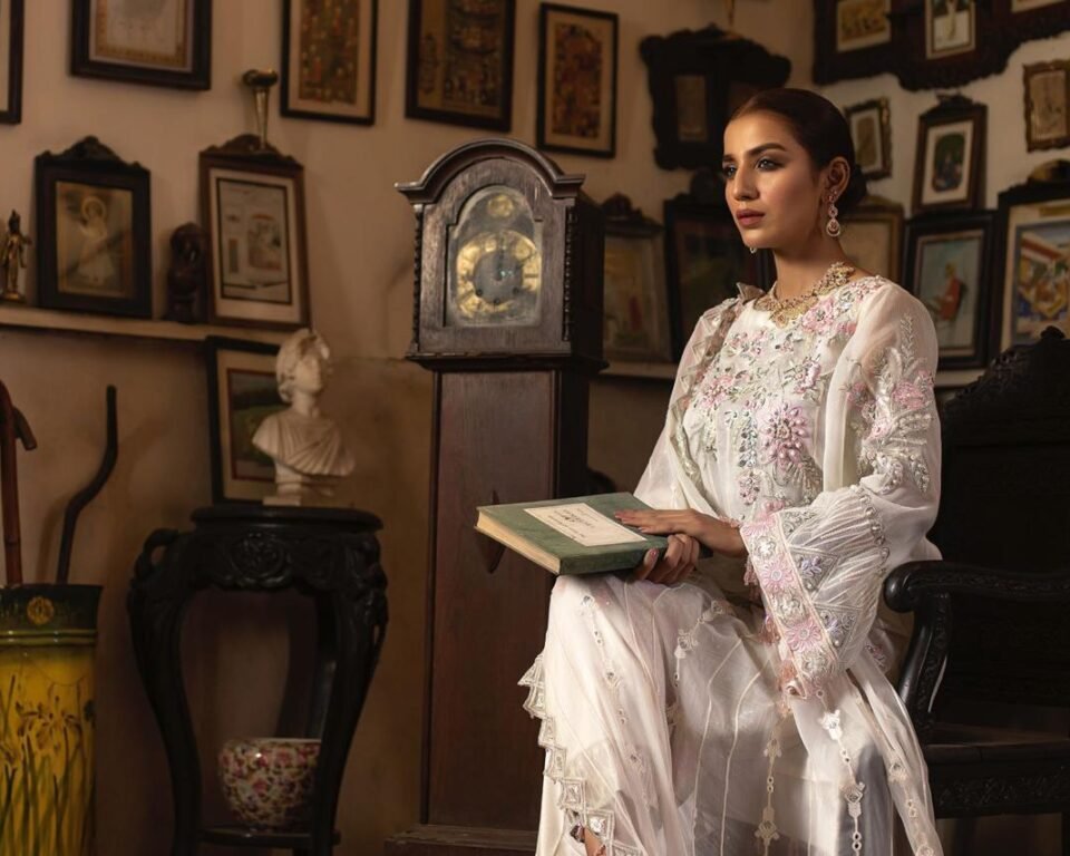 Editorial fashion photograph for the LUSH campaign featuring model Hafsa Janjua sitting indoors in a white embroidered traditional Pakistani outfit, holding a book, with antique frames and an old clock in the background. Fashion and cultural photography by AHSANZ.