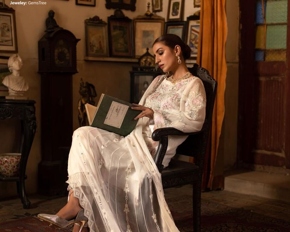 Vertical editorial fashion photograph for the LUSH campaign featuring model Hafsa Janjua sitting and reading a book in a white, embroidered traditional Pakistani outfit, surrounded by antique picture frames and a grandfather clock. Fashion and cultural photography by AHSANZ.