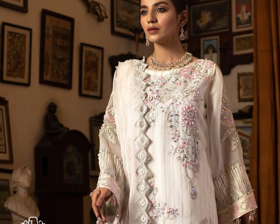 Close-up editorial fashion portrait for the LUSH campaign featuring model Hafsa Janjua standing, wearing a white traditional Pakistani outfit with intricate pink and green floral embroidery and a statement necklace. Wardrobe: LUSH, Jewelry: Gemstree. Fashion photography by AHSANZ.