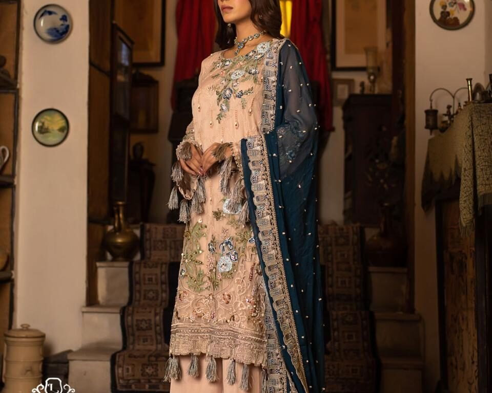 Full-length editorial fashion portrait for the LUSH campaign featuring model Hafsa Janjua standing in an antique room setting, wearing a blush-toned embroidered traditional Pakistani outfit with tassel details and a dark blue contrasting dupatta. Wardrobe: LUSH, Jewelry: Gemstree. Fashion photography by AHSANZ.