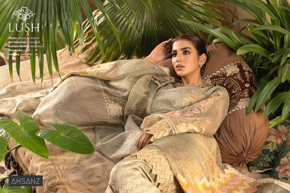 Horizontal editorial fashion photograph of model Hafsa Janjua reclining on patterned pillows in a lush, green outdoor setting, wearing a full, embroidered traditional Pakistani suit. Photo by Ahsan Ali Shahid.