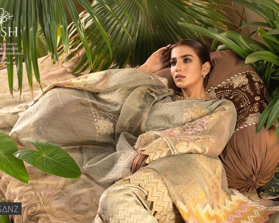 Horizontal editorial fashion photograph of model Hafsa Janjua reclining on patterned pillows in a lush, green outdoor setting, wearing a full, embroidered traditional Pakistani suit. Photo by Ahsan Ali Shahid.