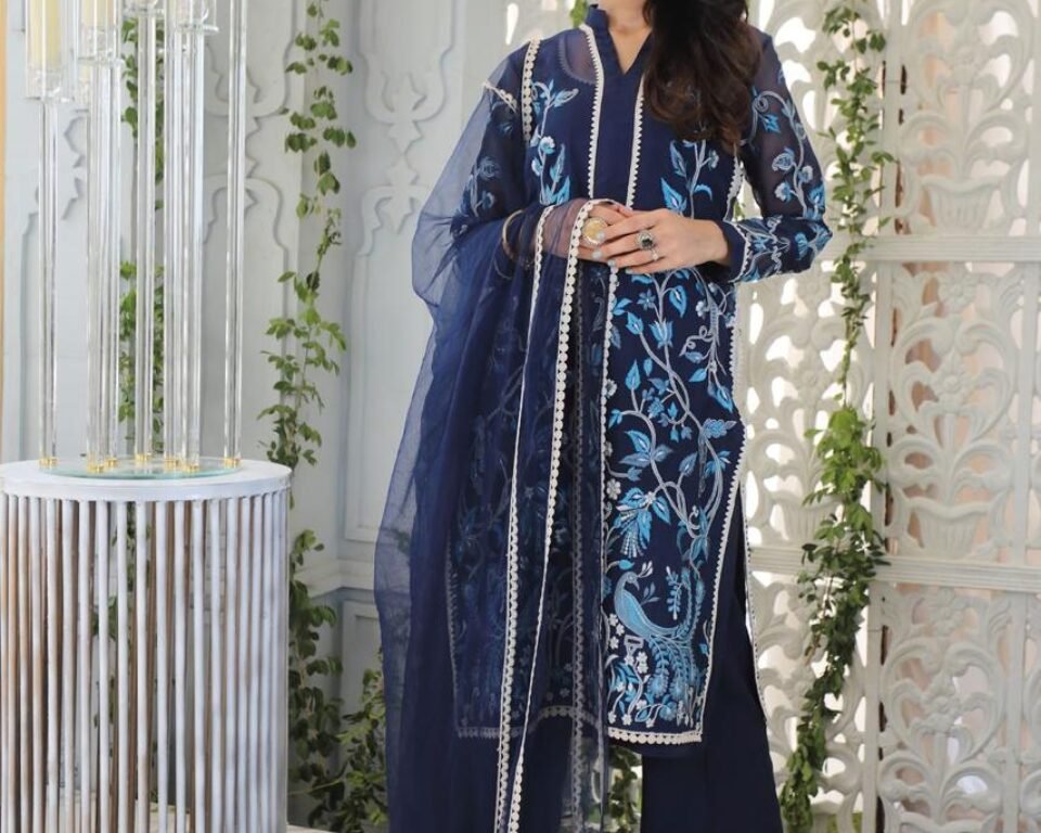 Full-length editorial fashion photograph of a model standing in a white, ornate room, wearing a dark navy blue traditional Pakistani suit with light blue and white floral and peacock-themed embroidery and a matching sheer dupatta. Fashion photography by AHSANZ.