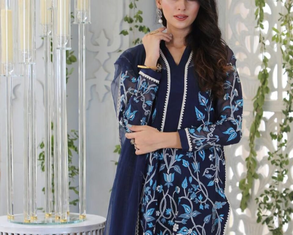 Close-up editorial fashion portrait of a model standing near a white decorative screen, wearing a navy blue traditional Pakistani suit with light blue and white floral and peacock-themed embroidery. Fashion photography by AHSANZ.