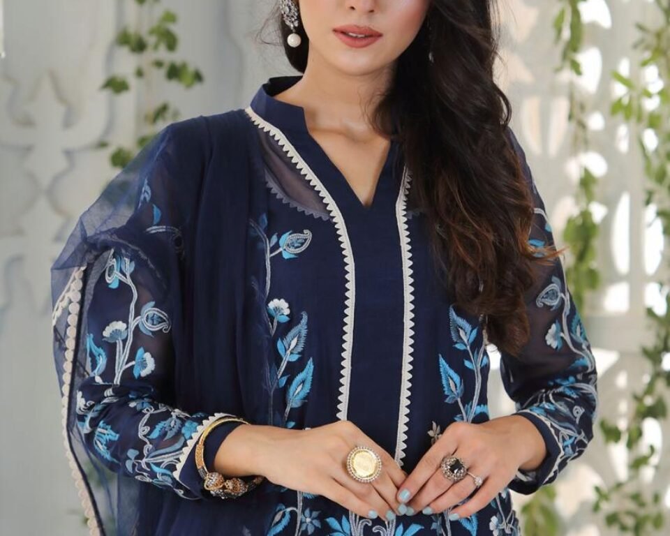 Close-up editorial fashion portrait of a model wearing a navy blue traditional Pakistani suit with light blue and white floral and peacock-themed embroidery and a matching sheer dupatta. The model is posing indoors with greenery decoration. Fashion photography by AHSANZ.