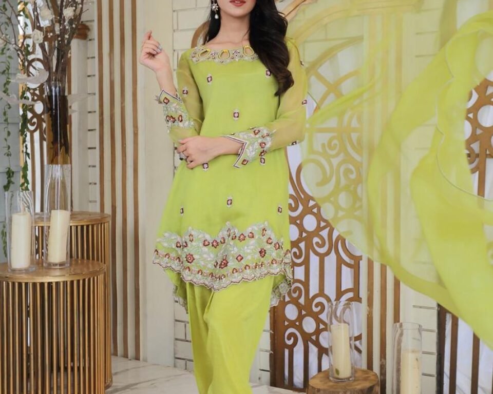Full-length editorial fashion photograph of a model wearing a sleeveless lime green high-low traditional Pakistani suit with red floral embroidery, paired with fitted trousers and gold heels, in a bright, stylized indoor setting. Fashion photography by AHSANZ.