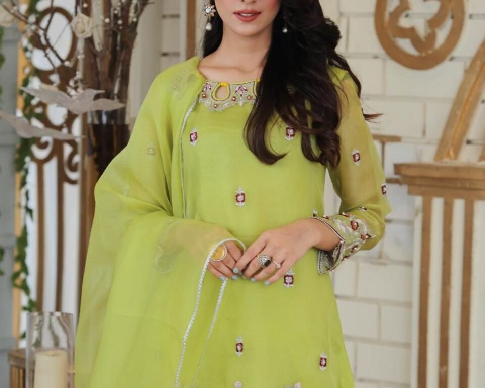 Close-up editorial fashion portrait of a model wearing a sheer lime green traditional Pakistani suit with red, white, and silver floral embroidery on the hem and sleeves, posing in a bright, stylized indoor setting. Fashion photography by AHSANZ.