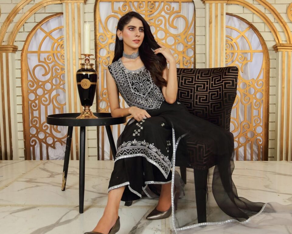 Editorial fashion photograph of a model sitting in a stylized room with a gold and white geometric backdrop, wearing a sleeveless black traditional Pakistani suit with heavy silver embroidery. Fashion photography by AHSANZ.