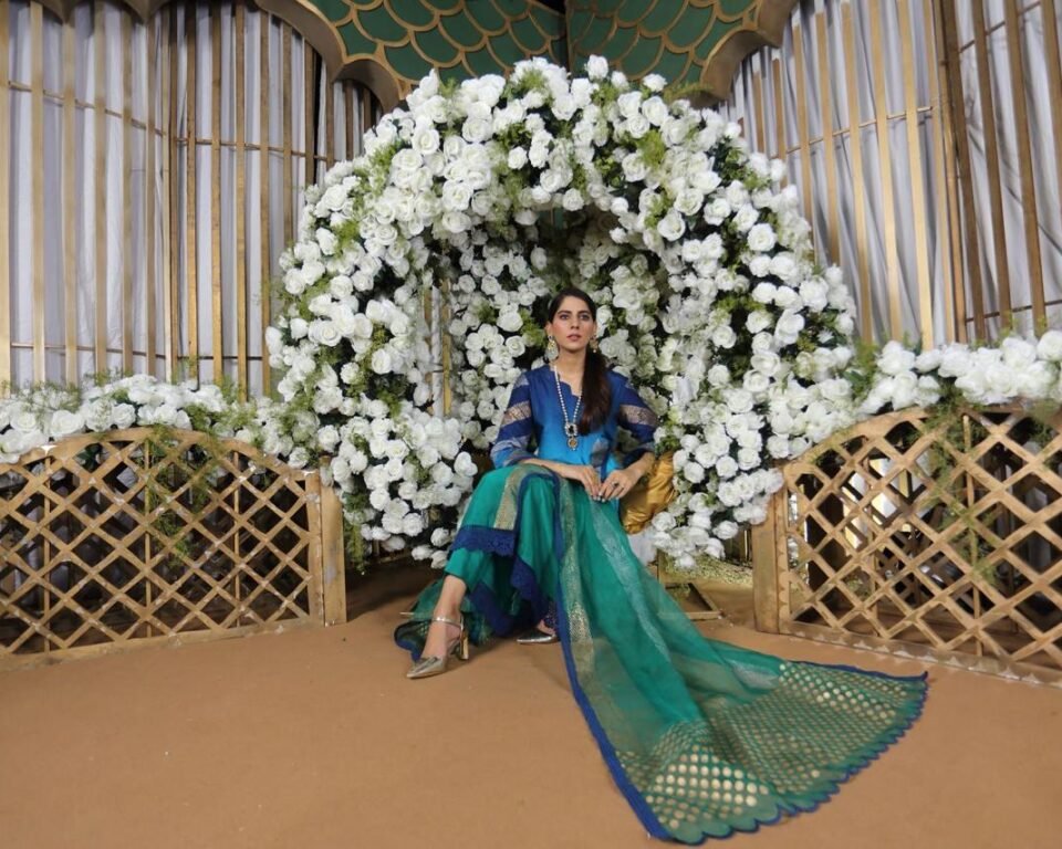 Editorial fashion photograph of a model sitting in a gold and green decorative archway covered in white roses, wearing a blue and green ombre traditional Pakistani suit with a flowing gold-dotted dupatta. Fashion photography by AHSANZ.