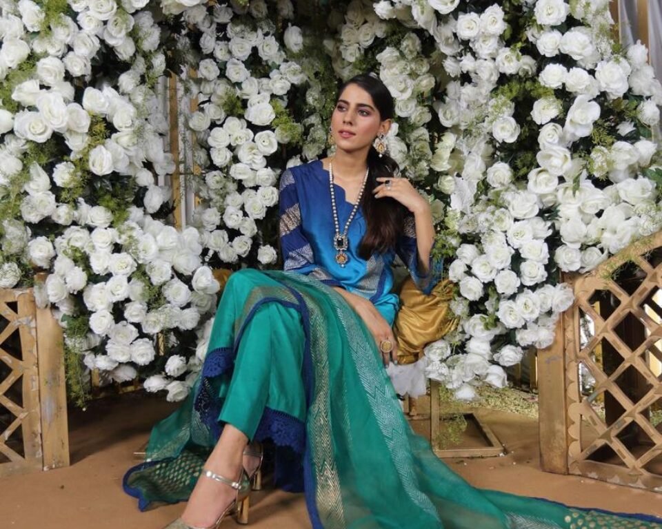 Full-length editorial fashion photograph of a model standing beneath an archway covered in white roses, wearing a dark blue and turquoise green ombre traditional Pakistani suit with a gold-dotted dupatta. Fashion photography by AHSANZ.