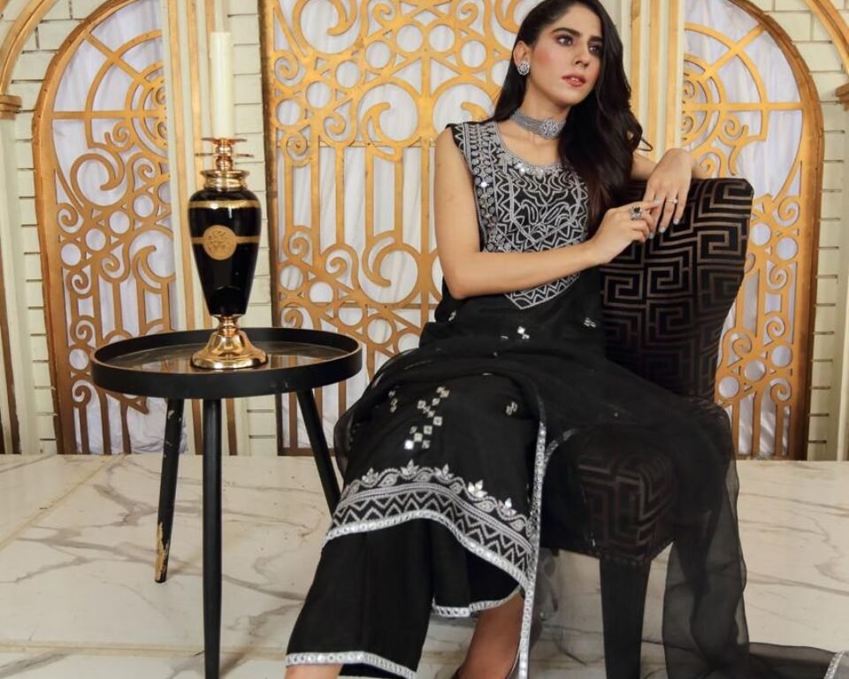 Full-length editorial fashion photograph of a model sitting in an ornate black velvet chair on a marble floor, wearing a black traditional Pakistani suit with heavy silver embroidery, set against a gold geometric backdrop. Fashion photography by AHSANZ.