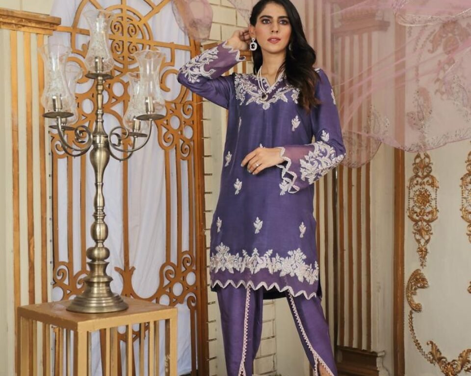 Full-length editorial fashion photograph of a model standing in a white and gold ornate room, wearing a purple traditional Pakistani suit with white floral embroidery, paired with tulip shalwar trousers. Fashion photography by AHSANZ.