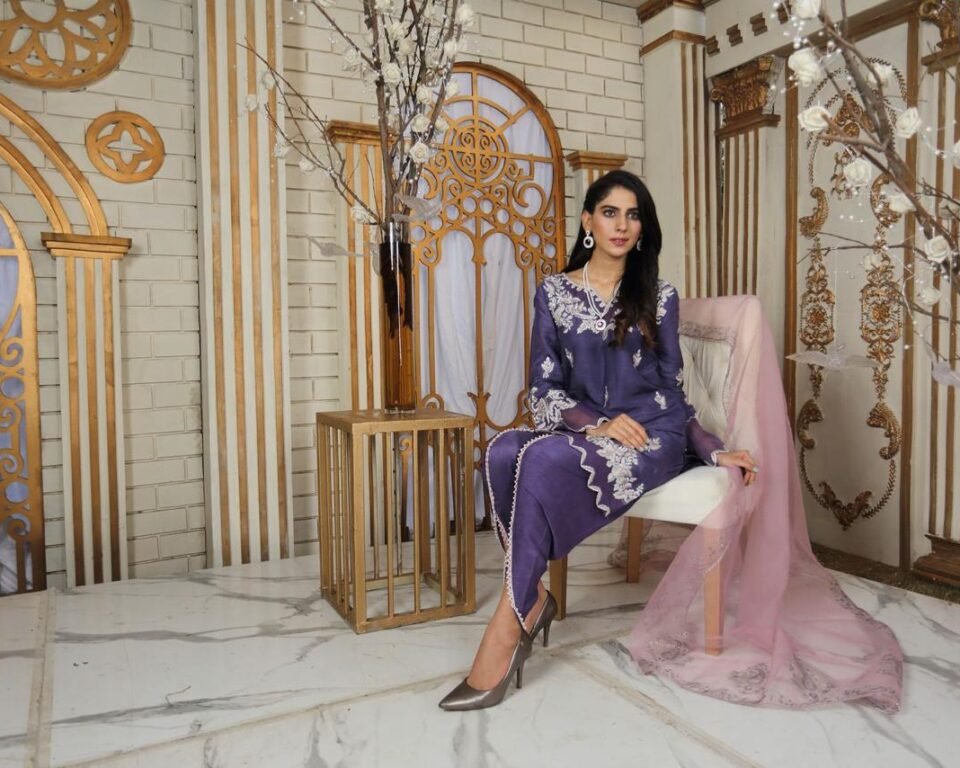Full-length editorial fashion photograph of a model sitting on a white chair in an ornate room with gold archways, wearing a purple traditional Pakistani suit with white embroidery and a pink sheer dupatta. Fashion photography by AHSANZ.