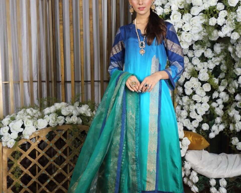 Full-length editorial fashion photograph of a model standing in a highly decorated room with white floral arrangements, wearing a dark blue and turquoise green ombre traditional Pakistani suit with gold and blue embroidery. Fashion photography by AHSANZ.