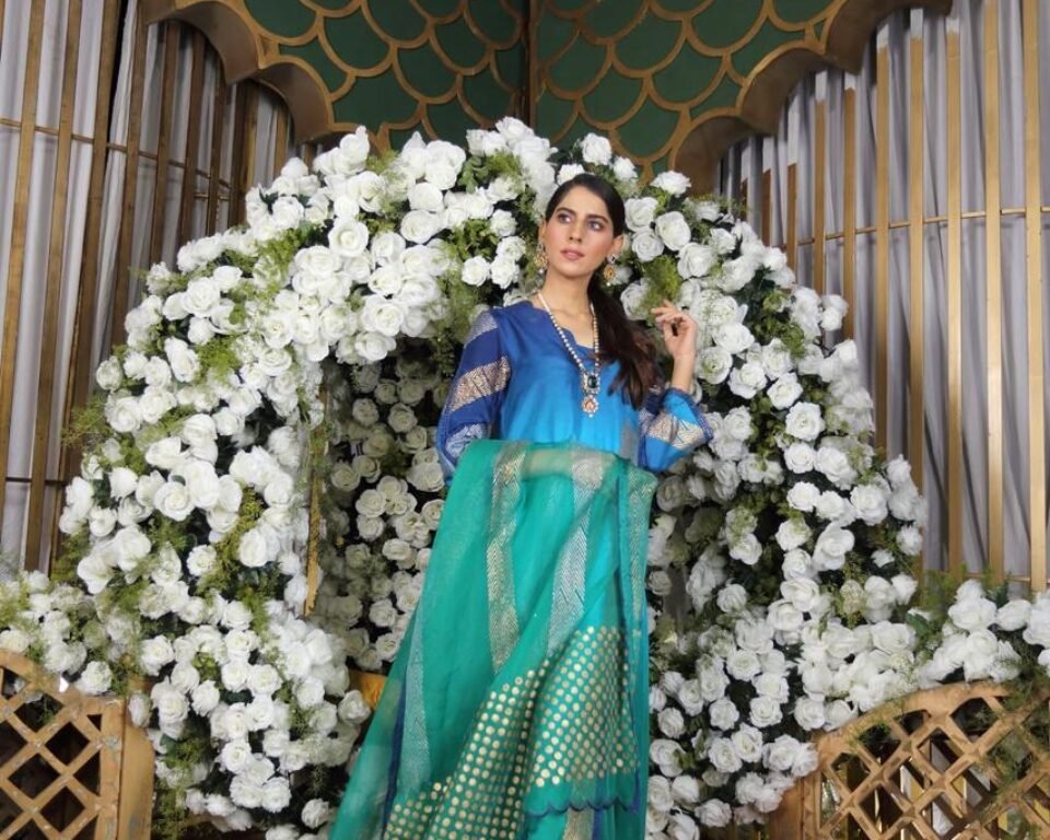 Full-length editorial fashion photograph of a model standing in a white and gold ornate room, wearing a purple traditional Pakistani suit with white floral embroidery, paired with fitted trousers and high heels. Fashion photography by AHSANZ.
