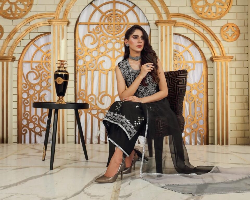 Close-up editorial fashion photograph of a model sitting against a white rose archway, wearing a dark blue and turquoise green ombre traditional Pakistani suit with a gold-dotted dupatta draped over the shoulder. Fashion photography by AHSANZ.
