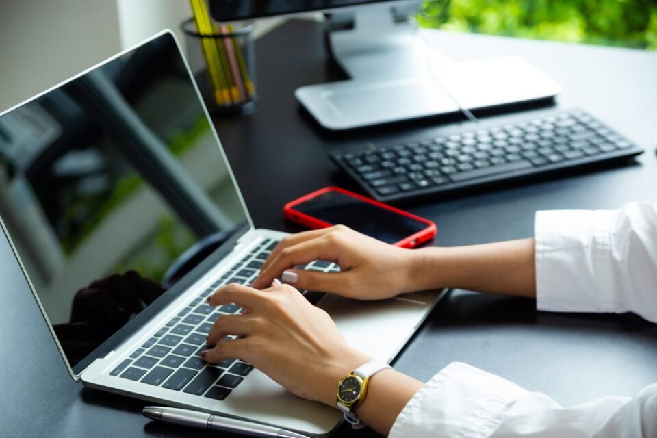Close-up photograph of a person in a white shirt typing on a silver laptop computer on a black desk, with a red mobile phone, desktop monitor, and keyboard visible in the background. Professional workspace photography asset by AHSANZ for website development services.